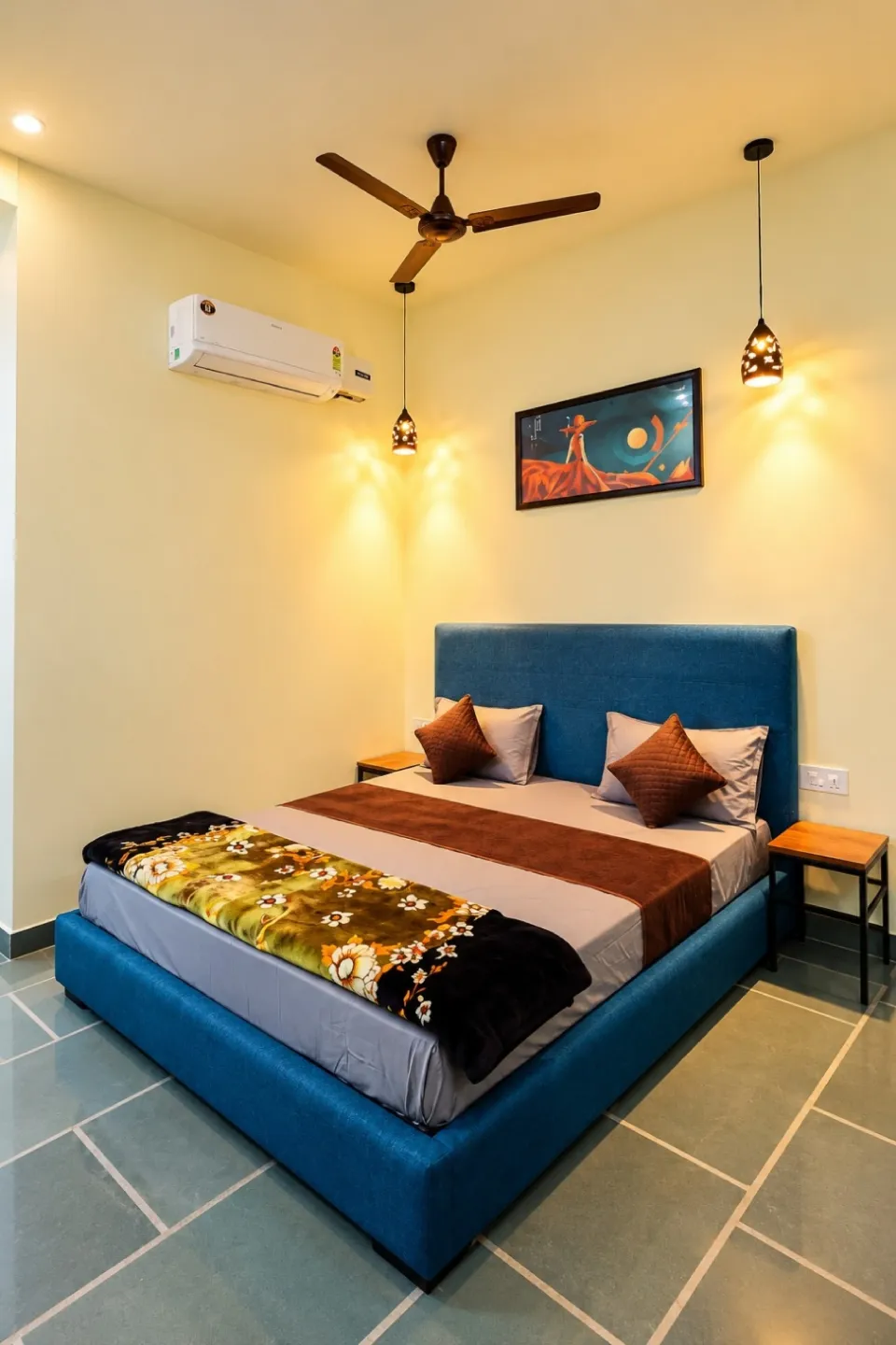 Teal upholstered bed with brown pillows, floral blanket, and warm pendant lights in a cozy bedroom