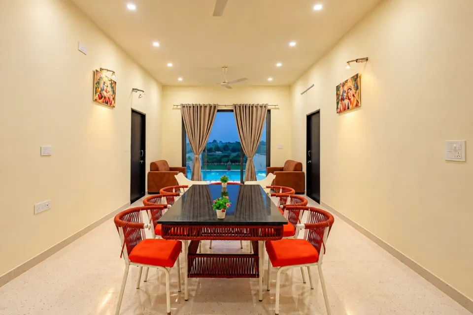 Spacious dining area with a black table, red cushioned wicker chairs, cream walls, and curtains overlooking a swimming pool