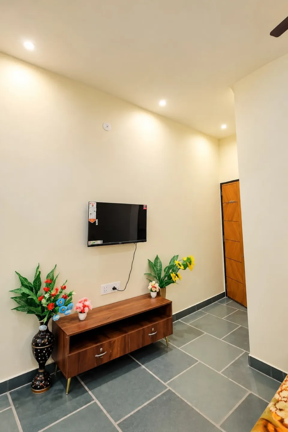 Modern room corner featuring a black TV, wooden TV cabinet, colorful artificial flowers, and a wooden door