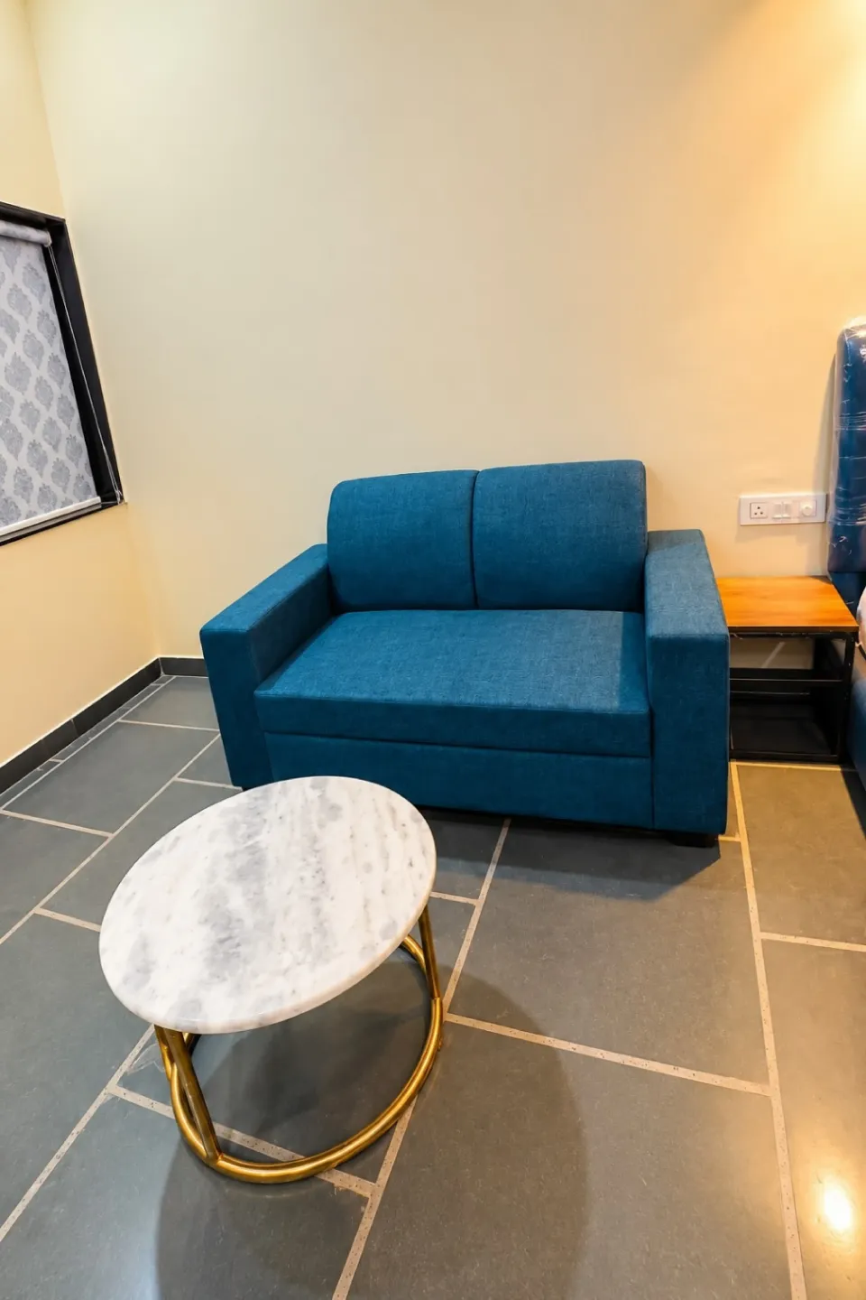 Modern room corner with a blue two-seat sofa, a white marble oval table, gold accents, and a small wooden side table