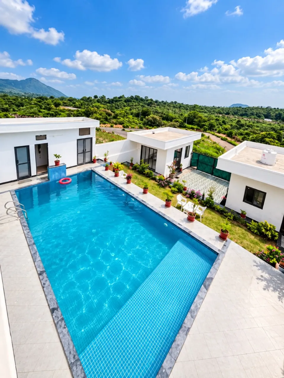 Outdoor swimming pool surrounded by modern white villas, potted plants, and lush green hills under a bright blue sky